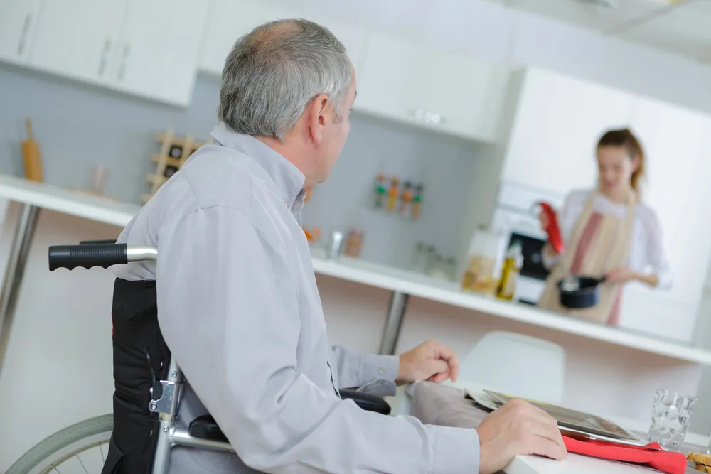 Man in a wheelchair in a kitchen while a caregiver cooks, representing independent living skills training