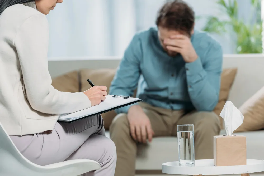 Therapist taking notes during a mental health treatment session with a distressed man